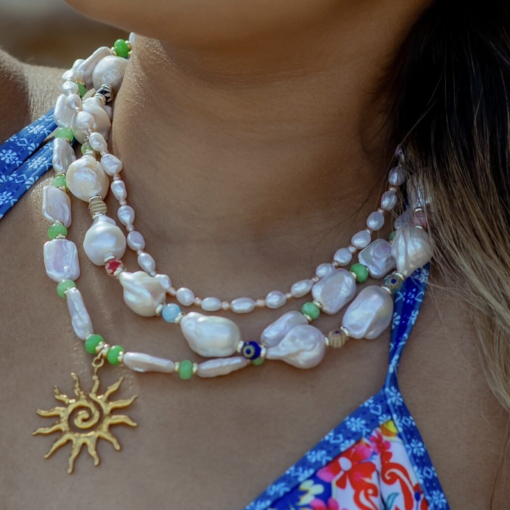 Close-up of layered pearl and bead necklace with a gold sun pendant resting on the chest of a person in a blue floral swimsuit top