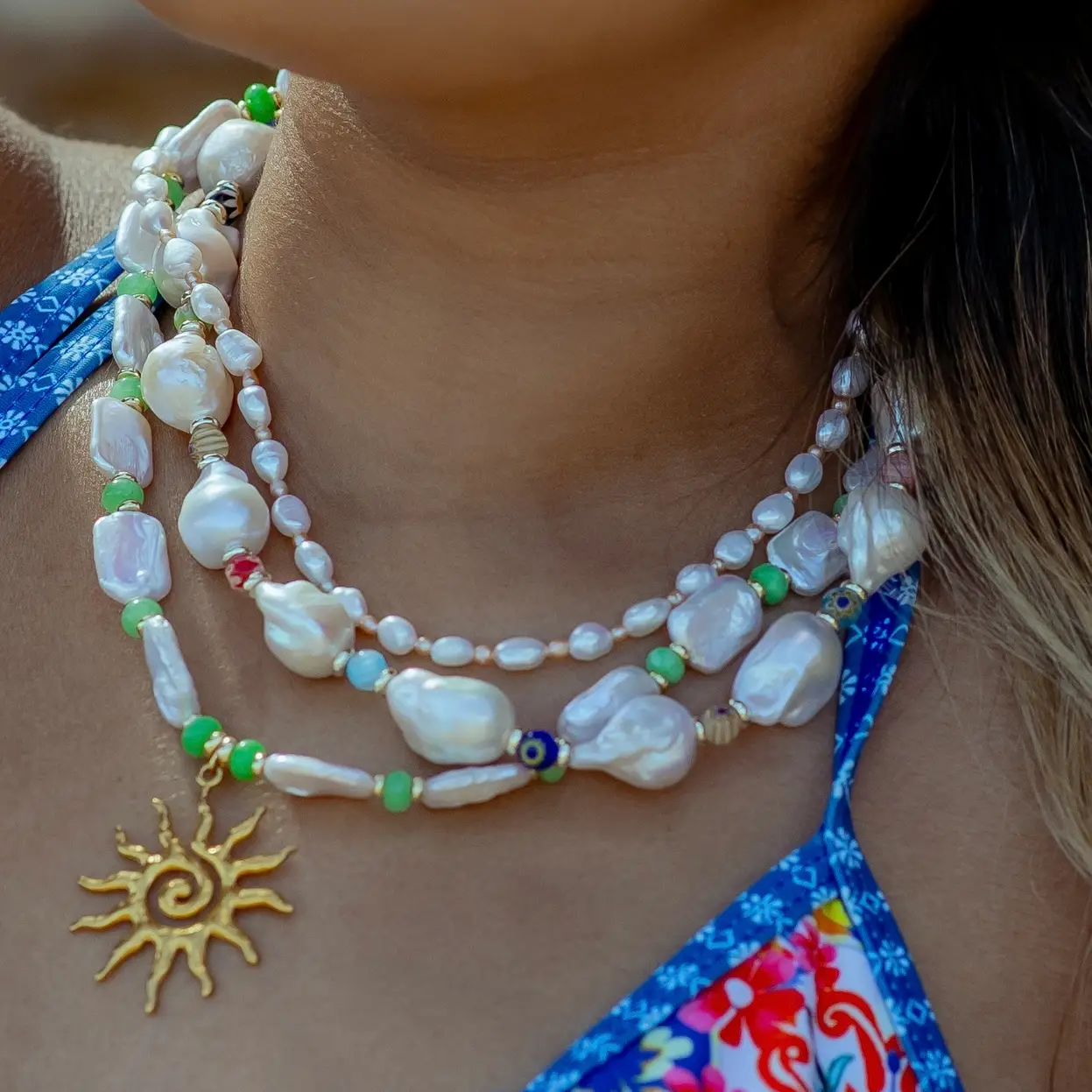Close-up of layered pearl and bead necklace with a gold sun pendant resting on the chest of a person in a blue floral swimsuit top