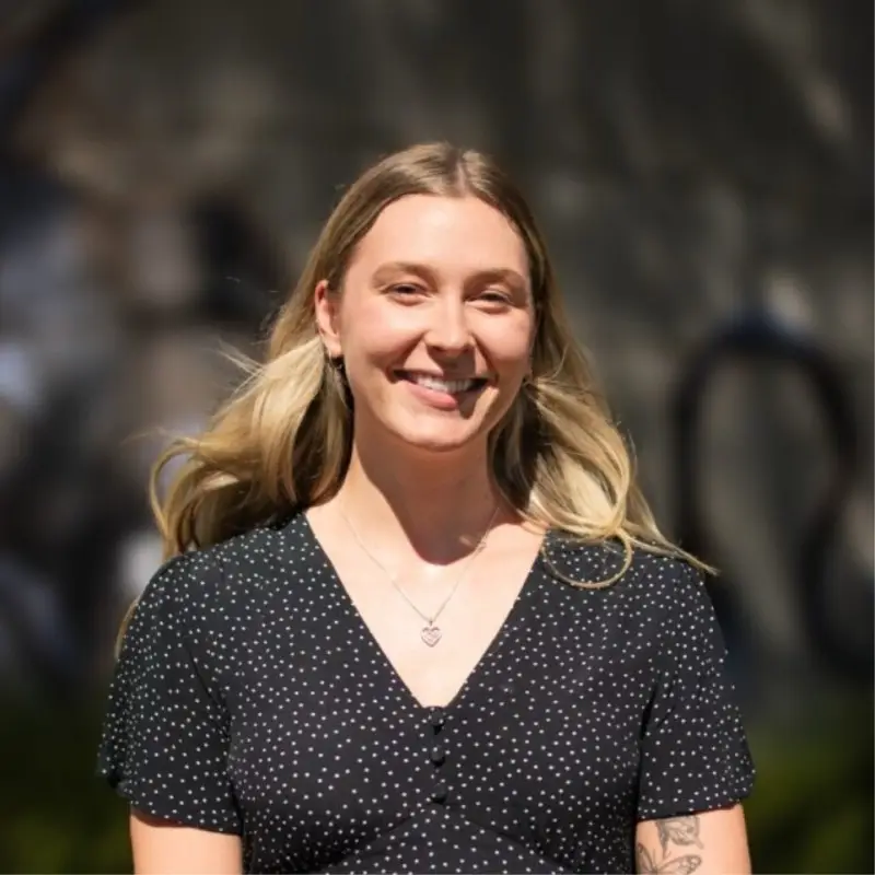 Smiling woman outdoors with long blonde hair, wearing a black polka-dot top and a heart pendant necklace.