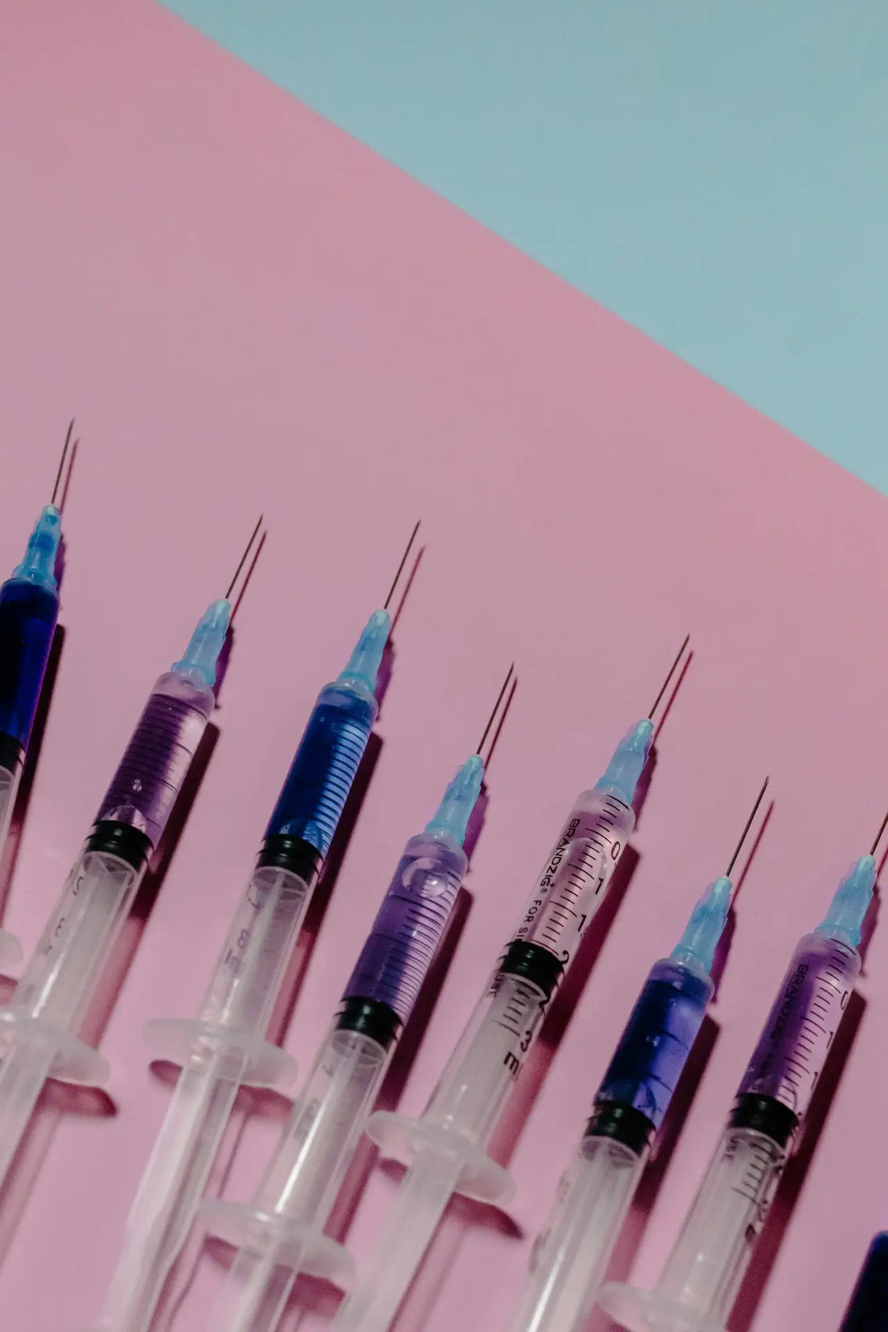 Row of syringes with colored liquids arranged diagonally on a pink background.