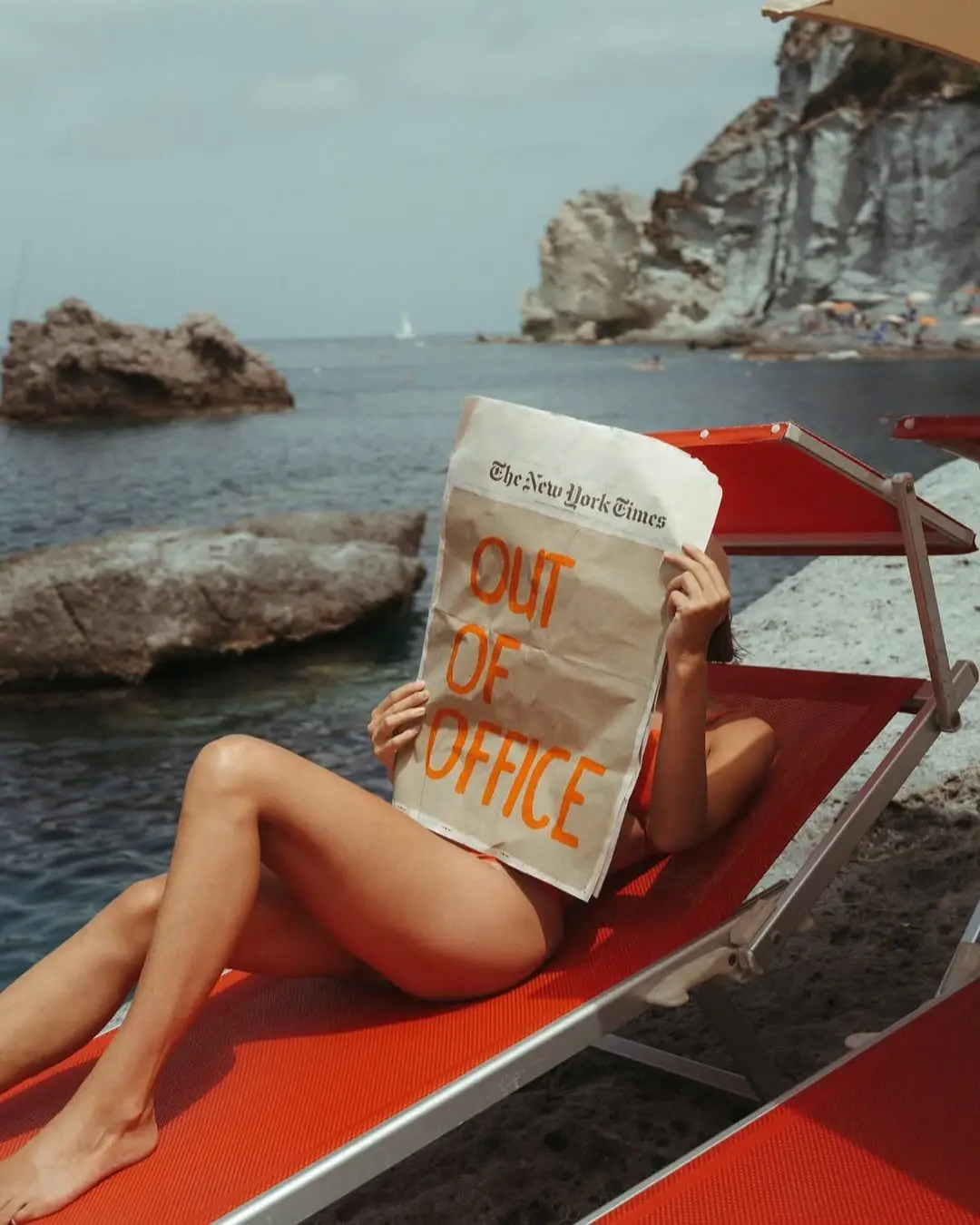 Person lounging on a red beach chair by the water, holding a newspaper with the headline 'OUT OF OFFICE' and 'The New York Times'.