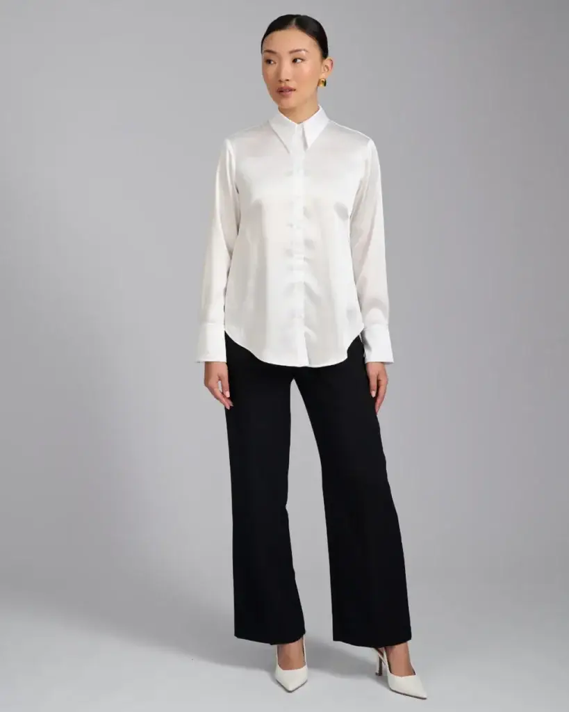 Woman in a white satin button-down blouse with black wide-leg pants and white heels, standing against a gray backdrop.