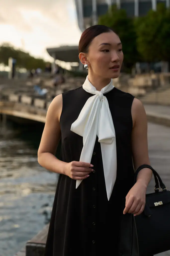 Portrait of a woman by the water wearing a black sleeveless dress with a white neck scarf, holding a black handbag.