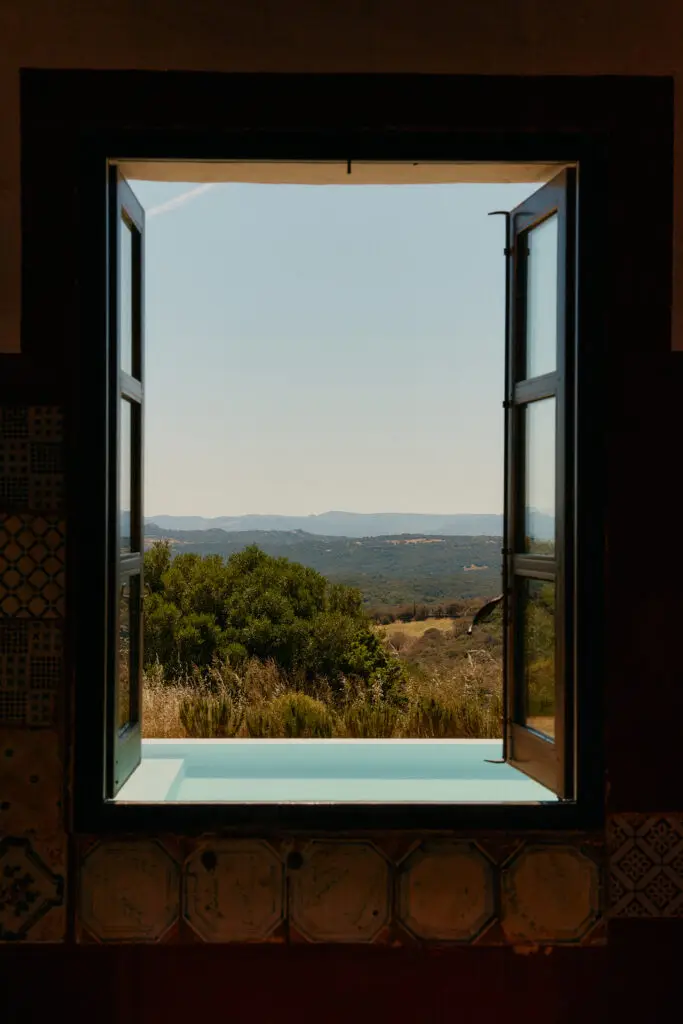 Open window frames a sunlit countryside with rolling hills and trees beyond