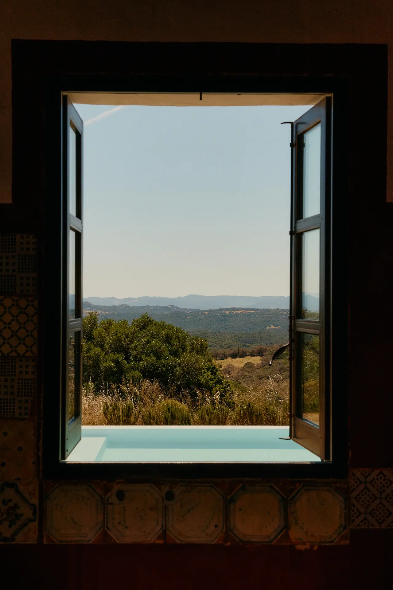 Open window frames a sunlit countryside with rolling hills and trees beyond