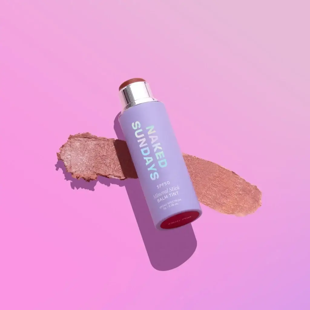 Lilac tube labeled 'NAKED SUNDAYS' angled on a bronze makeup smear against a pink background.