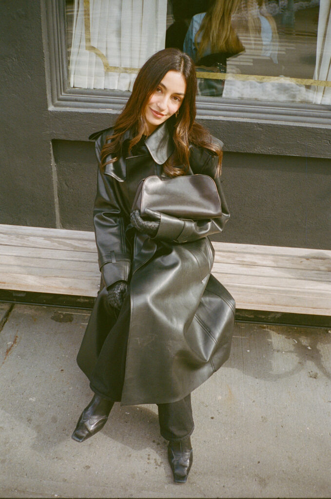 Woman in a long black leather coat sits on a wooden bench outside a storefront, holding a matching black bag.