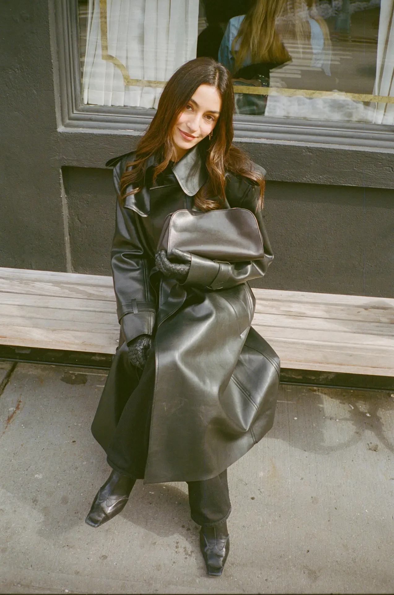 Woman in a long black leather coat sits on a wooden bench outside a storefront, holding a matching black bag.