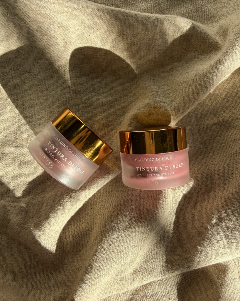 Two pink glass jars with shiny gold lids resting on beige fabric, labeled Tintura di Sole lip, cheek and eye tint.