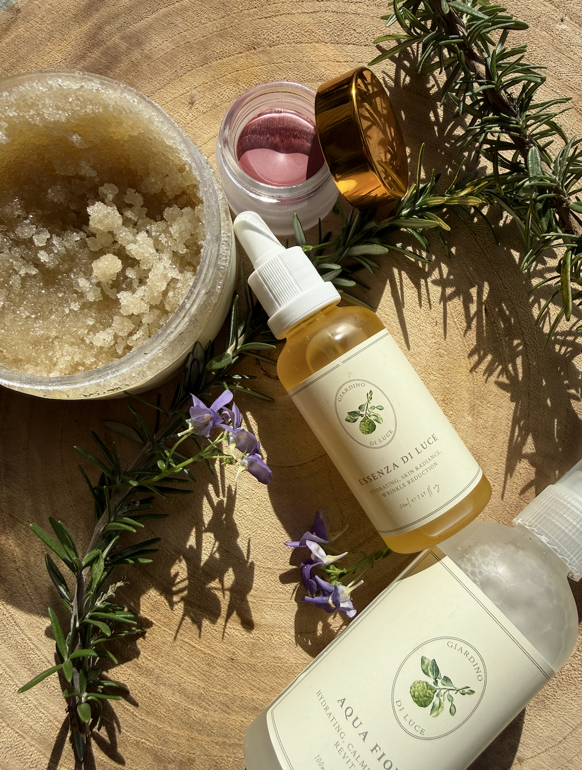 Oil and scrub skincare setup with a dropper bottle, jar of sugar scrub, and sprigs of rosemary on a wooden surface.