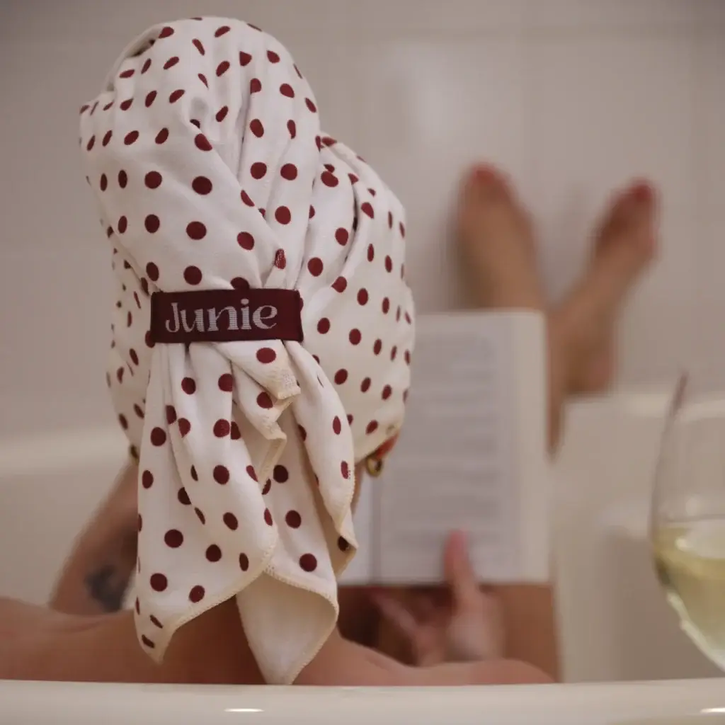 Person with a white towel dotted in red wrapped around their head, label 'Junie', in a bathroom near a tub mirror view
