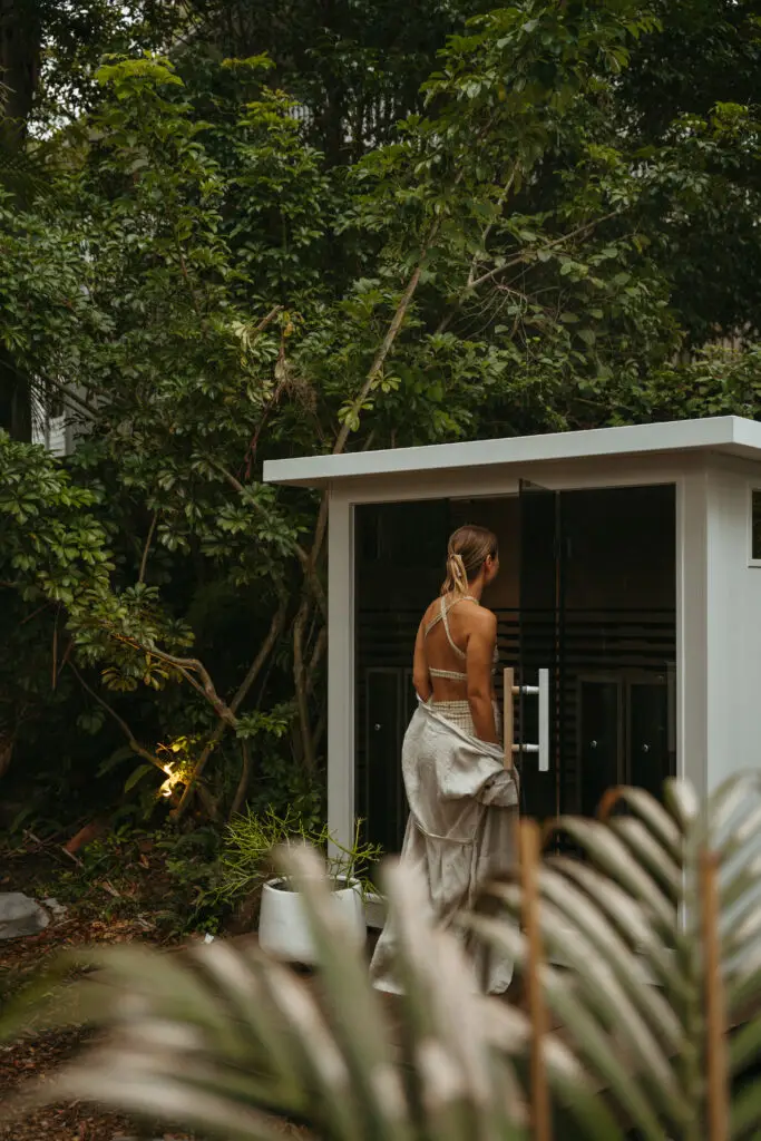 Woman seen from behind in a beige dress standing at a glass doorway in a lush garden area.