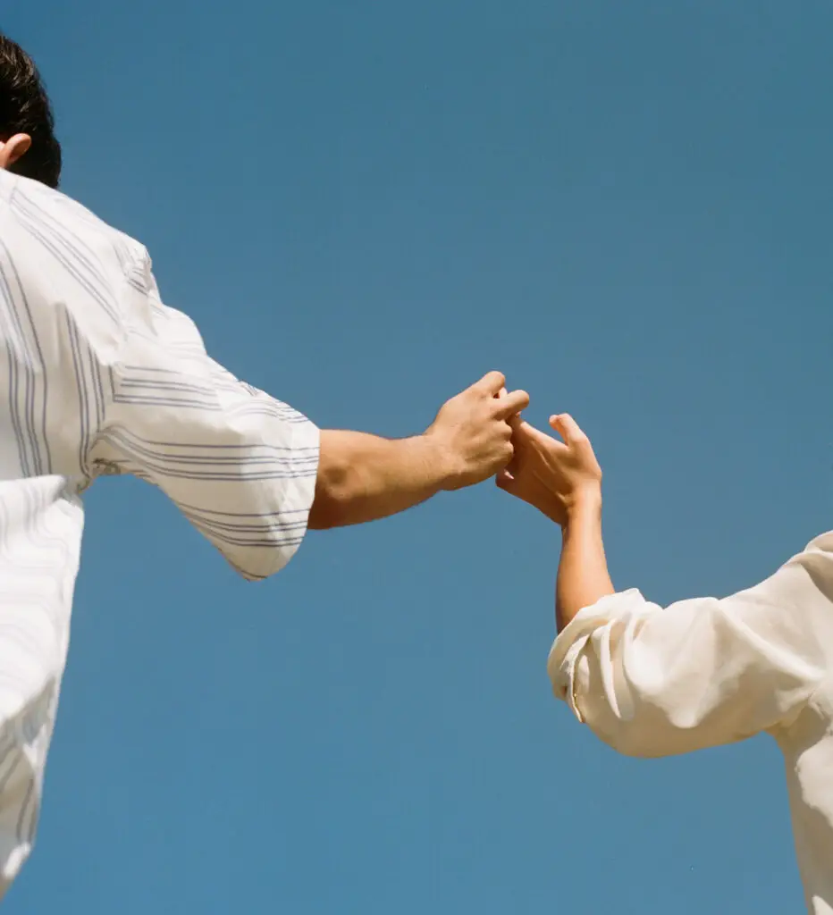 Two people reach toward each other and touch hands against a clear blue sky (outdoors).