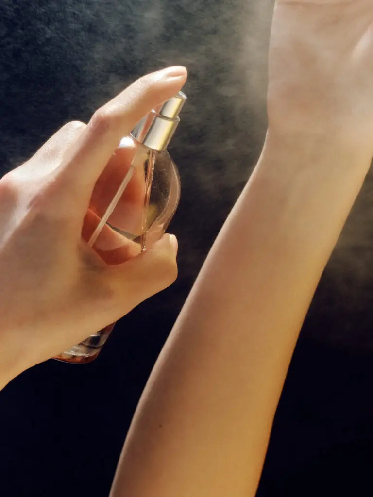 A hand holds a perfume bottle and spritzes fragrance onto the wrist of the other arm.