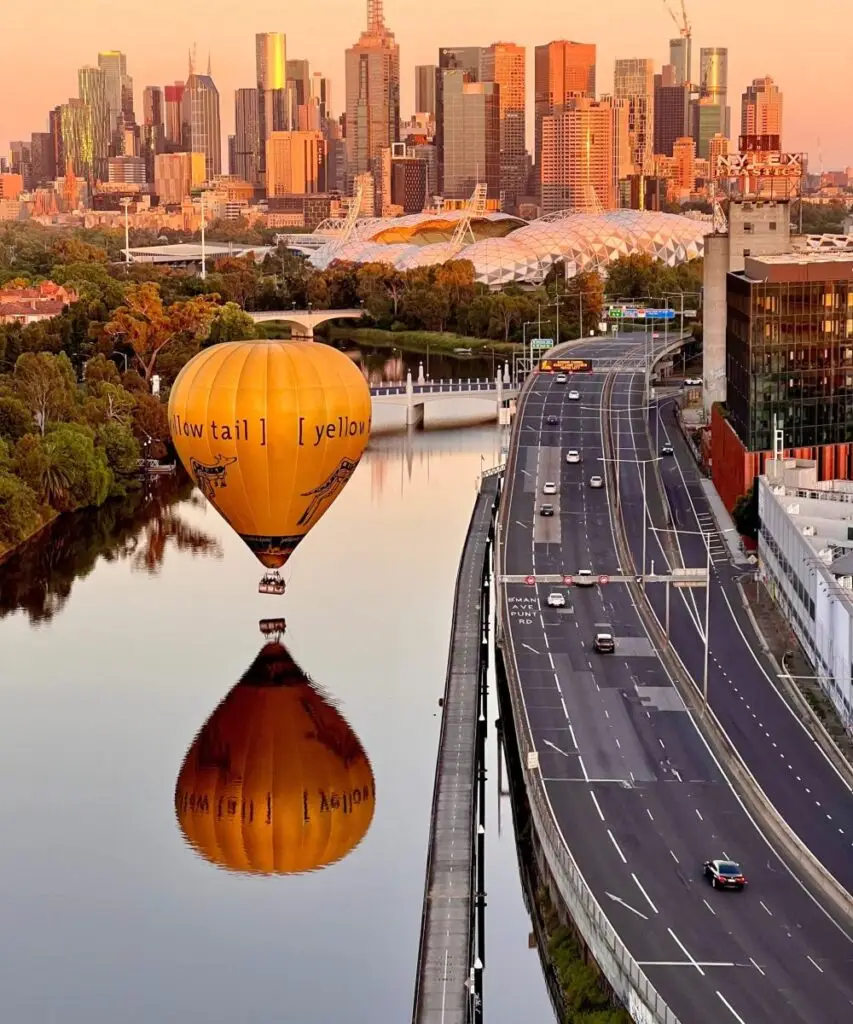 Orange hot air balloon over a calm river with Melbourne skyline at sunset; a multi-lane highway runs along the right side of the image, with reflections in the water.