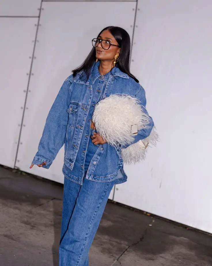 Woman in an oversized denim jacket and jeans, black glasses, gold earrings, holding a white fluffy bag outdoors.