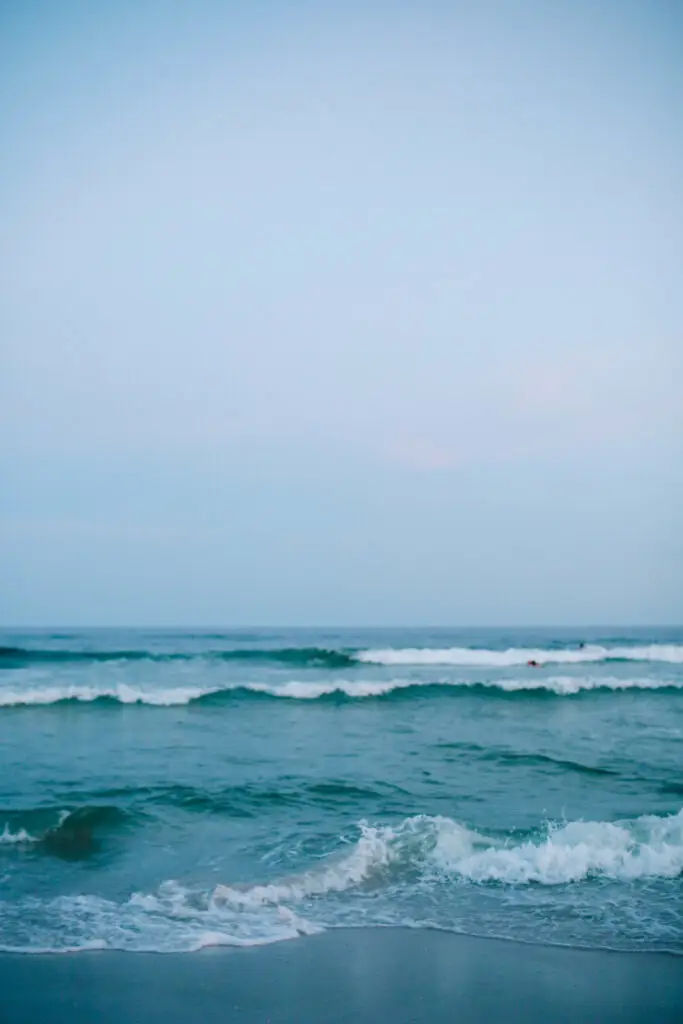 Ocean waves roll toward a sandy shore under a pale blue sky.