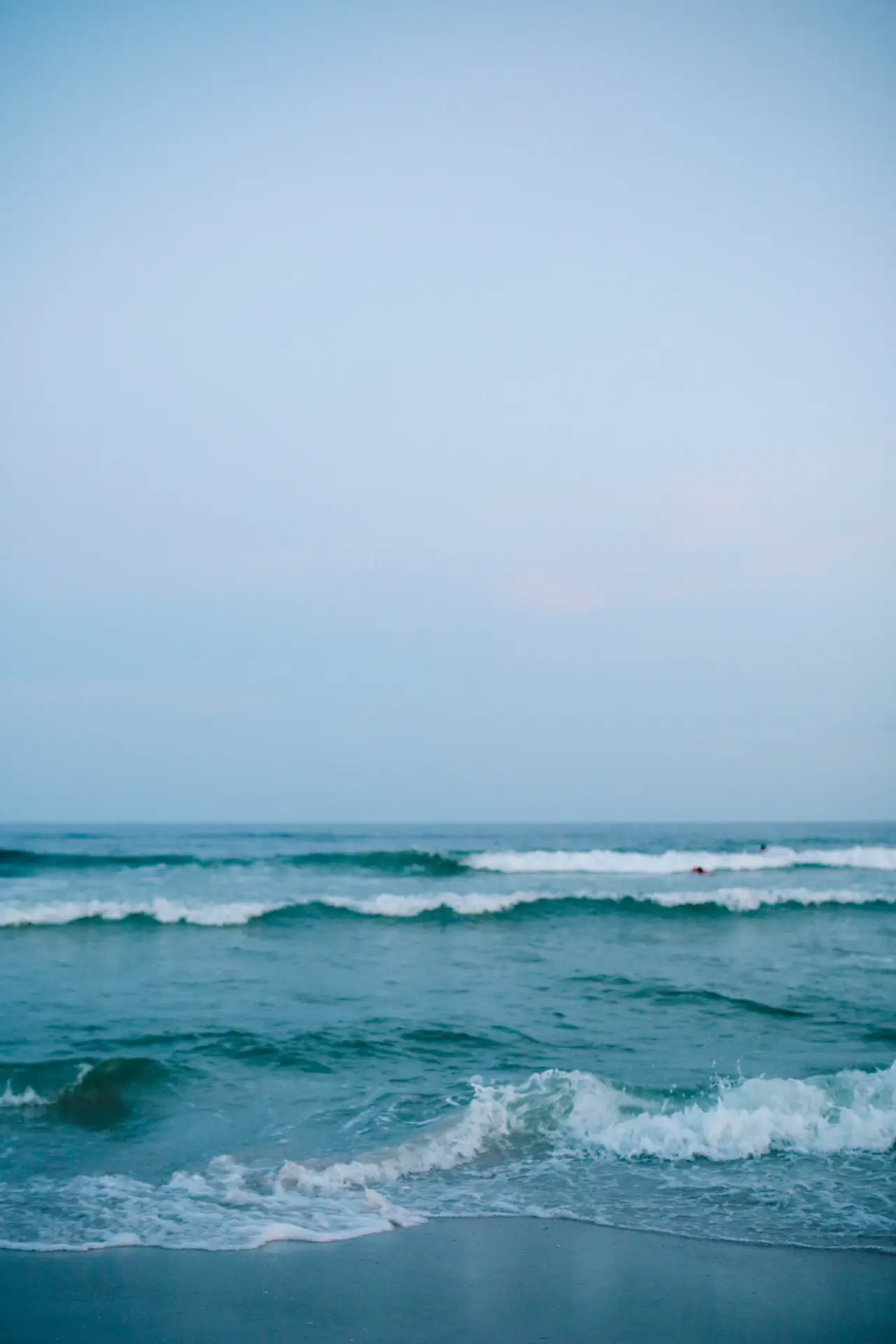 Ocean waves roll toward a sandy shore under a pale blue sky.