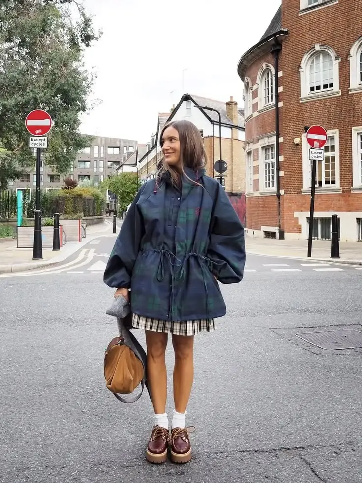 Woman standing at a street intersection wearing a navy plaid coat, plaid skirt, white socks, and brown platform shoes, carrying a brown bag and gray glove in her left hand.