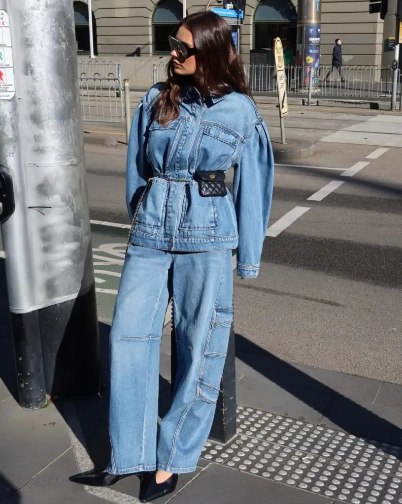 Woman wearing an oversized denim jacket and wide-leg jeans with a small black quilted belt bag, sunglasses, and black heeled shoes on a city sidewalk.